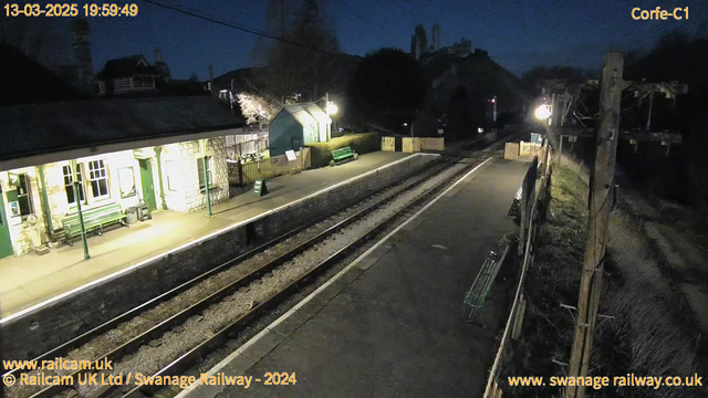 A dimly lit train station platform at twilight. The platform has two railway tracks and features a stone building with several windows. A green bench is visible near the entrance, with another green bench further down the platform. There are wooden fences along the edge of the platform, and a sign indicating "OUT" is placed near the building. Soft lighting illuminates parts of the platform, while shadows are cast in other areas. In the background, a hill is present, with some trees and structures partially visible.