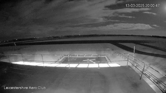 A dimly lit scene captured from a webcam shows a flat rooftop area with a fenced boundary. In the center, there is a white, illuminated section that outlines a cross shape, likely indicating a helipad. The background features a dark sky with scattered clouds, and a distant horizon where lights suggest the presence of nearby structures or roads. The image conveys a tranquil, nighttime atmosphere.