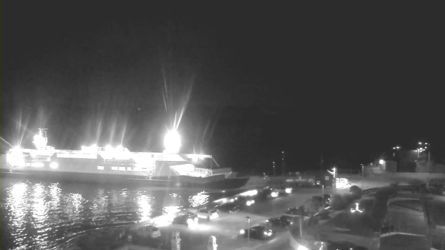 A brightly lit ferry is docked at a harbor at night, reflecting light off the calm water. The dock features several parked cars and illuminated pathways. In the background, a dark shoreline and distant lights can be seen against the night sky.