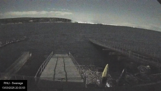 A dimly lit waterfront scene featuring a dark body of water with gentle waves. On the left, a wooden pier extends into the water, partly obscured by darkness. In the foreground is a concrete platform with a light-colored kayak resting against it. The background shows a distant rocky shoreline, illuminated slightly by a cloudy night sky.