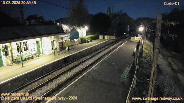 A nighttime scene at a railway station. The platform is illuminated with soft light, featuring a stone building with large windows, green benches, and a "Way Out" sign. Two railway tracks run parallel, with gravel between them. In the background, there are distant hills and trees, with a gentle slope leading to a hilltop. A single lamppost lights the right side of the image, and a wooden fence is visible along the platform edge. The atmosphere is quiet and serene, typical of a rural train station at night.