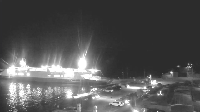 A bright ferry is docked beside a dark waterfront at night, illuminated by multiple lights that reflect on the water's surface. In the foreground, several parked cars are visible along a circular road, while the background features a faintly lit shoreline with some structures and streetlights. The scene conveys a quiet, nighttime atmosphere.