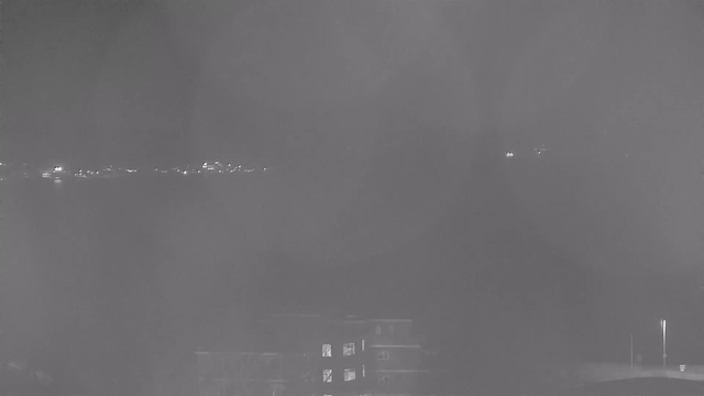 The image shows a dimly lit night view from a webcam, featuring a dark foreground with blurred shapes suggesting buildings. In the background, several small lights twinkle along the horizon, indicating the presence of distant structures or reflections on water. The overall tone is monochromatic with shades of gray, creating a foggy or hazy effect.