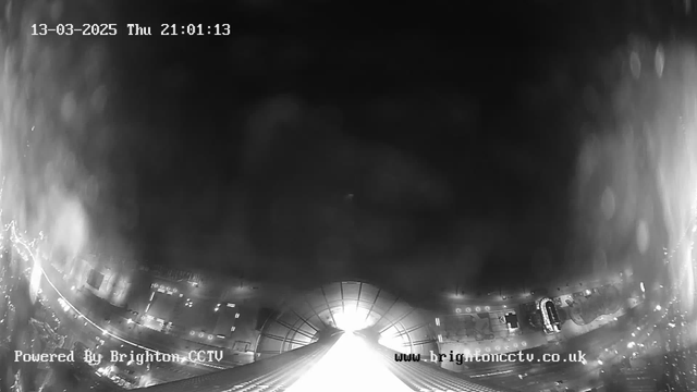 A black and white webcam image depicting a night scene. The center showcases a bright light source, while the edges reveal dark shapes suggesting a cityscape, possibly buildings or structures. The bottom of the image includes faint outlines of roads and vehicles with lights, creating a sense of urban activity. The timestamp in the upper left corner indicates the date and time, and the image is framed with a logo and web address at the bottom left and right corners.