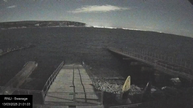 A nighttime view of a seaside area featuring a wooden pier extending into dark waters. There is a small boat ramp leading down to the water with several vertical lines visible on the surface. To the right, a yellow surfboard is propped upright on the ground next to some rocks. In the background, distant hills are partially illuminated by moonlight under a cloudy sky. The overall scene conveys a calm, quiet atmosphere.