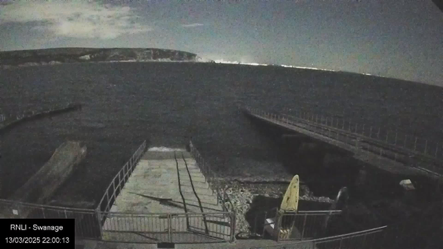 A dark, nighttime scene of a coastline, with a visible jetty extending into the water on the right side. The jetty is made of wooden planks and is lined with railings. In the foreground, there is a concrete ramp leading down to the water, bordered by fencing. Slightly to the left, a kayak lies on the ground. The water appears to have gentle waves, and in the background, a faint outline of a distant hill or cliff can be seen against the night sky, which has scattered clouds. A timestamp indicates the image was captured on March 13, 2025, at 10:00 PM.