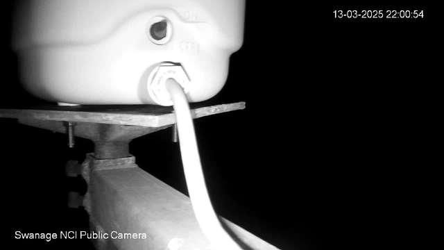 A close-up view of a white tank or container positioned on a metal support. The container has a visible outlet with a connecting white cable extending from it. The background is dark, making the details of the container and the support more prominent. In the bottom left corner, there is text indicating "Swanage NCI Public Camera." The date and time displayed in the top right corner are "13-03-2025 22:00:54."