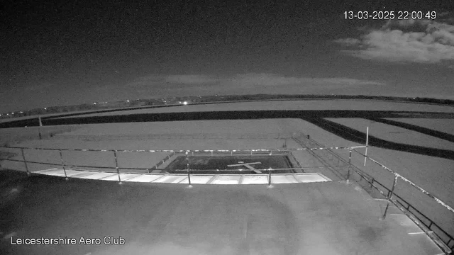 A nighttime view of a runway surrounded by a light railing. In the foreground, there is a helipad marked with a cross symbol. The sky displays some scattered clouds, and distant lights are visible along the horizon, indicating nearby structures or roads. The image is monochromatic, with varying shades of gray. The timestamp in the corner shows the date and time.