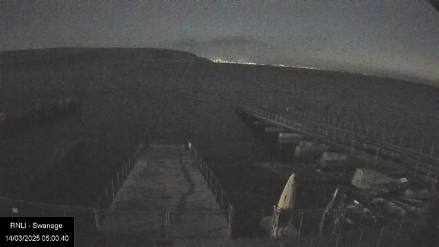 A dark, nighttime scene of a pier extending over water. The surface of the water is mostly calm, reflecting faint light from the horizon where a distant coastline is visible. The pier is lined with railing and leads toward a small cluster of boats at the edge. The atmosphere is dimly lit, with various shades of gray dominating the image.