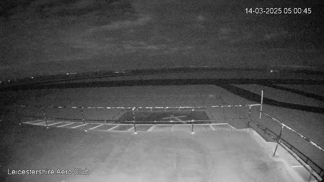 A low-light webcam image showing an empty tarmac area with a railing in the foreground. The scene is mostly dark with very faint outlines of a runway or parking area in the background. There are a few distant lights visible on the horizon, indicating possibly other buildings or vehicles far away. The sky is overcast with clouds. The timestamp in the top right corner indicates the date and time as March 14, 2025, at 5:00:45 AM. The name "Leicestershire Aero Club" is displayed in the bottom left corner.