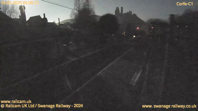 A dimly lit scene showing a railway track in the foreground with blurred outlines of buildings and trees in the background. The sky appears pale, suggestive of early dawn. There are faint lights indicating signals in the distance. The overall atmosphere is quiet and misty, suggesting a tranquil early morning setting.