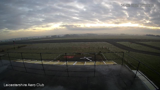 A view from a webcam at Leicestershire Aero Club captures a rural landscape at dawn. The foreground features a grassy area with a large white cross marked on the ground, near a building with railings. Beyond this, a runway is visible, bordered by a fence and patches of grass. The background shows a misty horizon and an overcast sky with varied gray clouds, indicating early morning light.