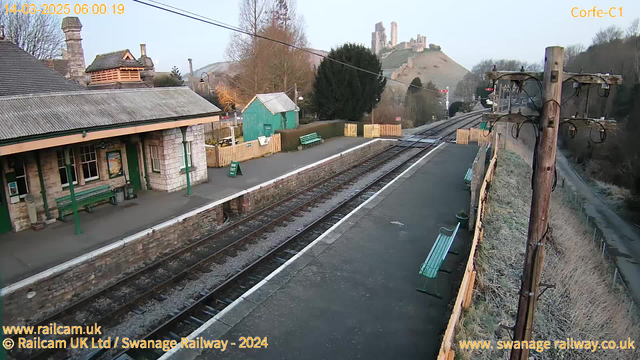 The image shows a railway station platform early in the morning. It features a stone building with a gabled roof and large windows on the left, alongside green benches and a sign indicating "WAY OUT." Further along the platform, there is a wooden fence and additional green benches. Tracks run parallel to the platform, leading towards the horizon. In the background, a hill rises, topped with a castle ruin. The scene is illuminated by the soft morning light, with trees visible in the surroundings. A telephone pole with several wires stands at the right edge of the image.