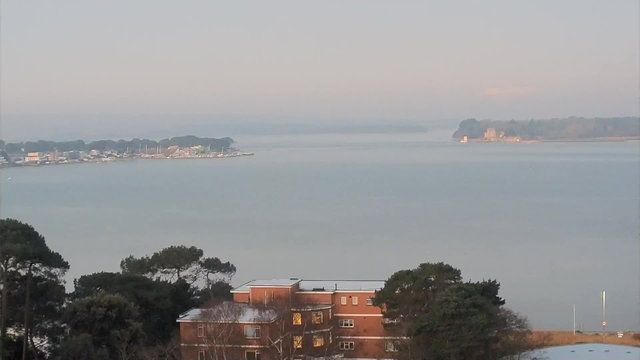 A calm waterfront scene captured from a webcam. In the foreground, there is a building with multiple windows and a flat roof, surrounded by trees. The mid-ground features a body of water reflecting soft light, with a small marina and a few boats visible along the shoreline. The background shows rolling land covered in trees, with a distant area that appears elevated, possibly a hill or a cliff. The sky is light, creating a serene atmosphere.