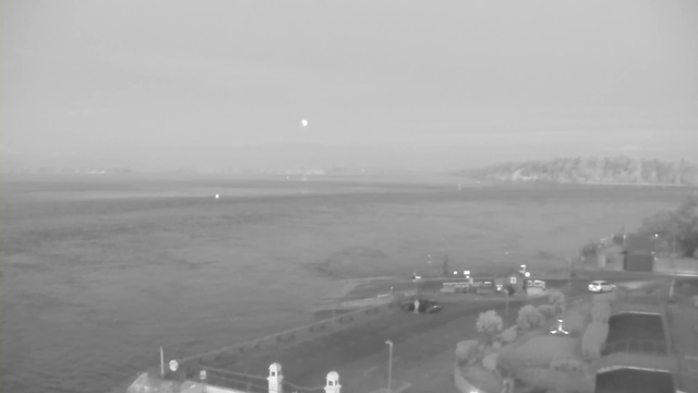 A grayscale image of a waterfront scene taken from a webcam. In the foreground, there is a paved area with some landscaping, including shrubs and a small fence. To the left, there are white pillars marking the entrance to the area. The midground features calm water with a few small boats visible. In the background, a distant landscape of hills can be seen, partially shrouded in mist. The sky appears overcast, giving a muted and tranquil atmosphere.