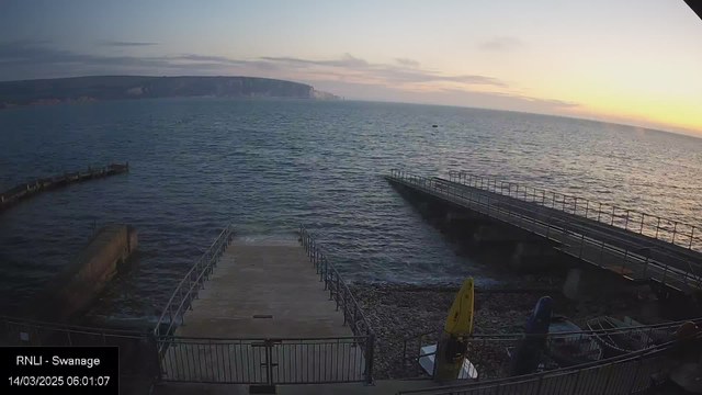 A seaside view at dawn, featuring calm blue water and a rocky shoreline. In the foreground, a wide staircase leads down to the water's edge, flanked by metal railings. To the left, a curved pier extends into the water, while to the right, a flat, straight pier reaches out further. There are several small boats stored near the base of the stairs and a yellow kayak on the shore. In the distance, a low cliff line sparsely decorated with greenery contrasts against the soft colors of the early morning sky, which transitions from pale blue to warm hues near the horizon.