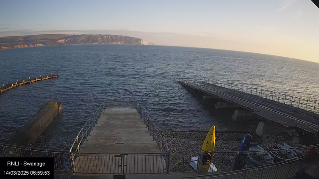 A calm seascape at dawn featuring a rocky shoreline. In the foreground, there is a concrete ramp leading down to the water, flanked by railings. To the right, there's a wooden pier extending over the water. Several kayaks are stored on the shore, with one yellow kayak prominently displayed. The background shows gentle waves and distant cliffs illuminated by the early morning light, under a clear sky.