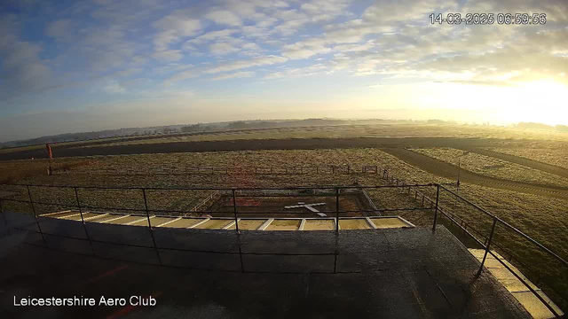 A view from a webcam at the Leicestershire Aero Club shows a grassy area and an airstrip under a bright sky with scattered clouds. In the foreground is a metal railing leading down to the airstrip, which is partially covered in grass and has markings for landing. The scene is illuminated by early morning sunlight, casting a warm glow across the landscape.