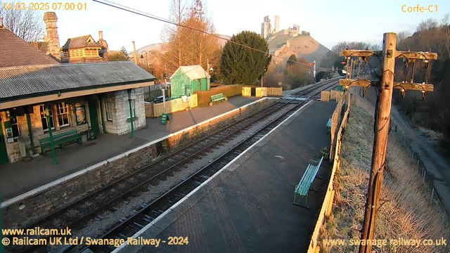 A view of Corfe Castle Railway Station at dawn. The foreground shows a railway platform with a green bench and a stone wall. In the background, there is a small green shed and a tree. The railway tracks run alongside the platform, leading into the distance. A large hill with Corfe Castle ruins is visible in the background, partially covered by trees. The scene is illuminated by soft morning light, creating a peaceful atmosphere.