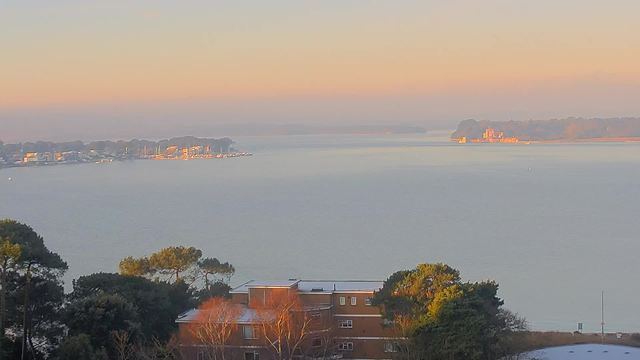 A scenic view of a calm body of water under a pastel sky at dawn. On the left side, a marina is visible with several boats docked. The coastline is lined with trees and buildings. In the foreground, there are some trees and a few buildings, while the horizon showcases hazy landforms in the distance.