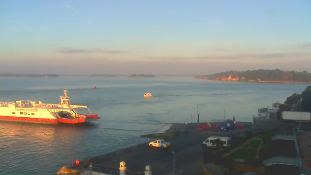 A tranquil waterfront scene showing a large white and red ferry docked, with a smaller orange boat navigating the water nearby. The water is calm and reflects the soft morning light, while a gentle mist hangs in the distance. Along the shoreline, there are green trees and a glimpse of a well-kept area with vehicles parked, including a white van and some red objects. The sky is clear with a soft blue hue, indicating early morning.