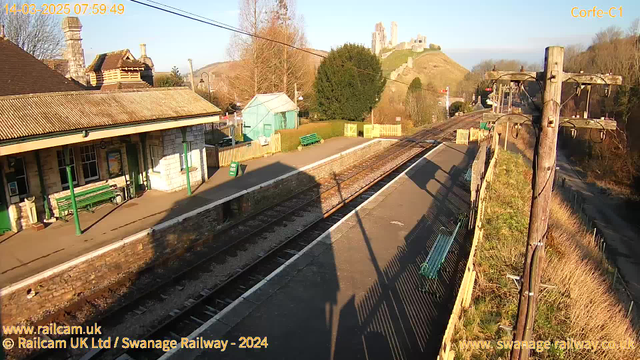 A sunny morning at Corfe Castle train station. The foreground features a railway platform with metal benches and a wooden fence on the right. There are two sets of railway tracks leading into the distance. On the left, a stone building with a sloped roof houses ticket booths and seating areas. Behind the station, a green hill rises, topped with the ruins of Corfe Castle. Trees are scattered around, and the sky is clear with a hint of blue. A sign that reads "WAY OUT" is visible on the platform.