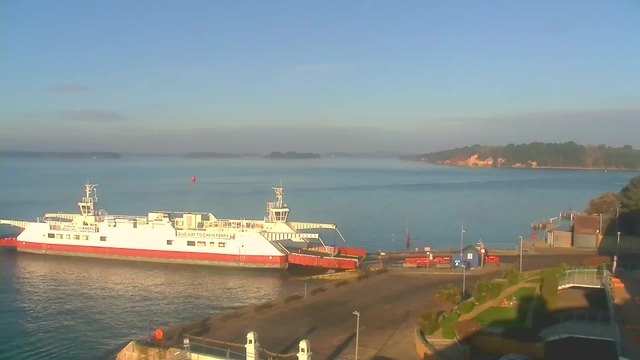 A ferry docked at a harbor sits calm in the water under a clear blue sky. The ferry is partially turned towards the left side, showing a white and red color scheme, with a ramp extended down to the pier. In the background, a distant shore with trees and a gentle slope is visible, while the water reflects the morning light. Several docks and boats are situated along the shore, with bright orange safety barriers and a small building nearby, all cast in soft shadows.