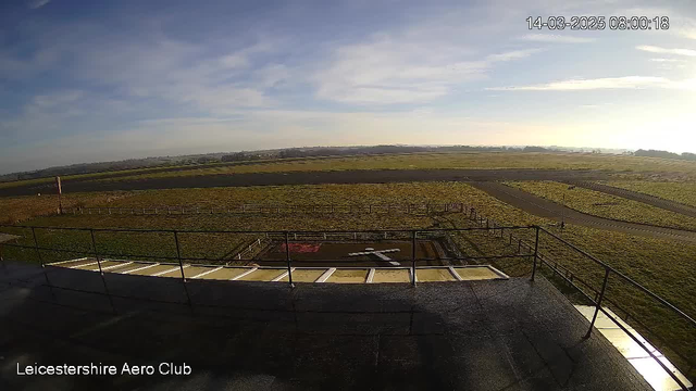 A webcam image shows a flat, expansive landscape under a clear blue sky. In the foreground, there is a gray rooftop railing, with a section of pavement visible. Below, an open field is visible, marked by a cross shape made from white material on the ground. A short runway or taxiway appears to the right, with patches of grass surrounding the area. There are some distant hills on the horizon and a few scattered clouds in the sky. The time and date are displayed in the top right corner.