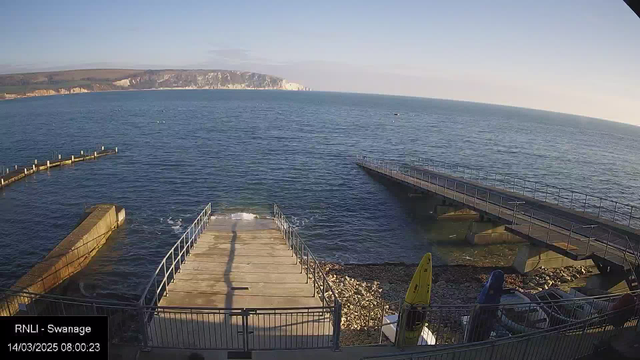 A panoramic view of a calm seaside scene featuring a wide expanse of water under a clear blue sky. In the foreground, there is a wooden jetty extending over the water, leading to a small concrete platform. To the right, two additional jetties stretch into the water, while a pile of stones and some kayaks in bright colors are visible along the shore. In the background, white cliffs rise above the water, creating a scenic natural backdrop. The timestamp indicates it is early morning.