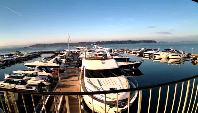 A scenic waterfront view featuring a marina filled with numerous boats and yachts docked along a wooden pier. The water is calm, reflecting the blue sky and distant shoreline. In the background, gentle hills are visible along the horizon. The scene conveys a serene and peaceful atmosphere, with clear weather and ample sunlight.