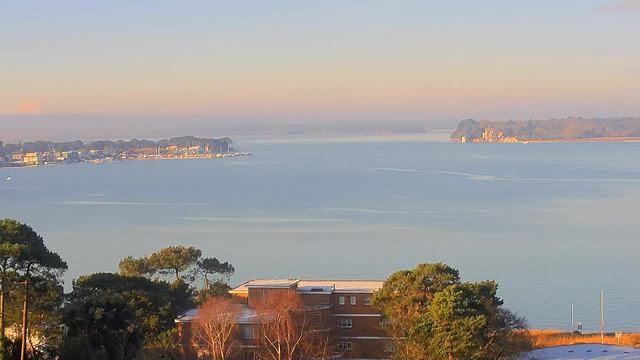 A serene view of a calm body of water reflecting soft hues of blue and pink, suggesting early morning light. In the distance, a small harbor lined with boats and buildings is visible against a backdrop of lush green trees. A few structures are situated in the foreground, partially obscured by greenery, while the water gently ripples. The sky appears clear with a pastel color gradient, enhancing the tranquil atmosphere of the scene.