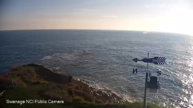 A coastal scene overlooking the ocean on a clear day. The water appears calm and shimmering in the sunlight, which is positioned in the upper right corner of the image. In the foreground, there is green grass and rocky terrain leading to the water's edge. A weather vane is visible on the right, indicating the directions North and South. The horizon stretches across the image, blending the sea and sky.