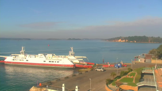 A white ferry with red accents is approaching a dock on the left side of the image, surrounded by calm blue water. In the background, there are distant green shores and trees. The sky is clear with a few clouds, and the scene is well-lit. Near the dock, there are vehicles parked and a landscaped area with grass and small palm trees.