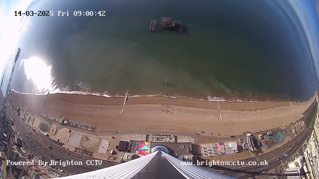 A wide aerial view over a sandy beach with gentle waves lapping at the shore. The water is calm and reflects sunlight, creating a glistening effect. There are a few people walking along the beach, and a long pier extends into the water, visible in the distance. In the foreground, colorful geometric patterns on a structure can be seen, along with various beachfront establishments. The image captures a clear blue sky with a few wispy clouds.