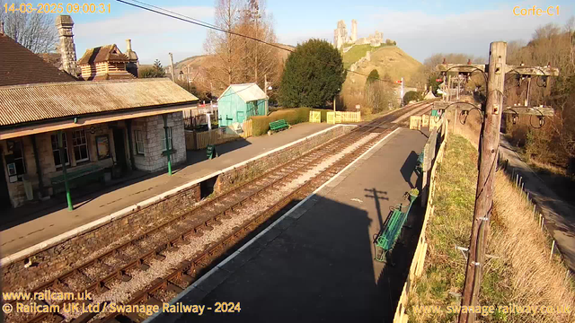 A railway station platform is shown on a clear sunny day. The foreground features tracks separated by gravel and a paved path leading to a stone building with a green balcony. Several benches are placed along the platform. In the background, a ruin of a castle is visible on a hill, surrounded by trees and grass. The sky is bright blue with a few scattered clouds. A wooden pole with wires stands on the right side of the image.