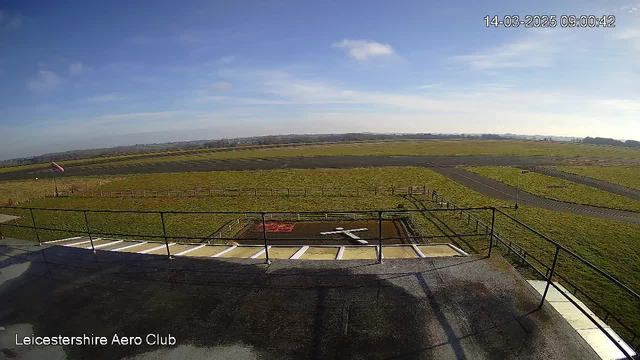 A panoramic view of an airfield with a grassy landscape. In the foreground, there is a railing and a small area marked with a white cross on the ground. The sky is mostly clear with a few clouds. To the left, a flag is visible, possibly indicating the wind direction. The airfield is bordered by a low fence, and the ground is mostly green with some darker patches. The scene conveys a calm and open setting with no visible aircraft.