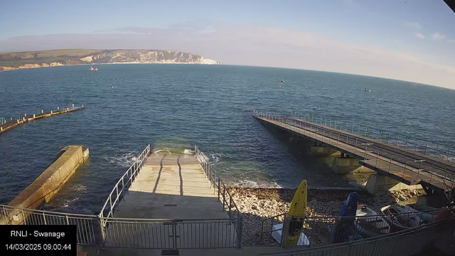 A view of a harbor with calm blue water and a rocky shoreline. In the foreground, there are wooden docks extending into the water, one straight and the other curving to the left. A yellow kayak is visible on the beach, along with other small boats. The background features white coastal cliffs under a clear sky. The time stamp at the bottom indicates it is 9:00 AM on March 14, 2025.