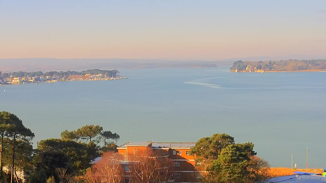 A panoramic view of a calm body of water under a clear sky. In the foreground, there are trees and buildings partially obscured by vegetation. The water stretches into the distance, where small boats are visible. On the far shore, a marina with multiple sailboats and a coastal village can be seen, along with a line of trees and a faint outline of hills in the background. The overall atmosphere is serene and bright, suggesting a peaceful day.