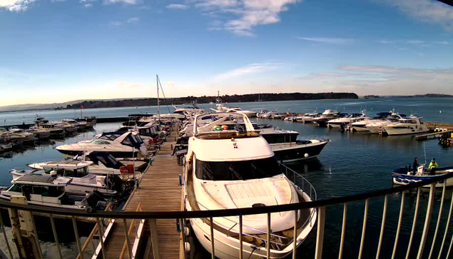 A marina scene with numerous boats docked along a wooden pier. Some boats are large yachts, with a prominent white boat in the foreground. The background shows a calm blue sea under a partly cloudy sky. There are hills visible in the distance. The area appears tranquil and sunny.