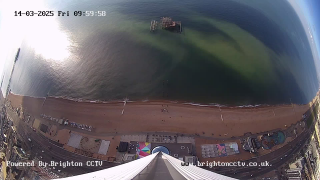 A high-angle view of a coastal area with sandy beaches and a pier in Brighton. The image shows the sea with a shimmering surface, reflecting sunlight. There is an old, partially submerged pier in the water. The beach is lined with various structures including colorful beach huts and an amusement area visible along the shore. The scene is captured under clear skies, giving a bright and vibrant atmosphere.