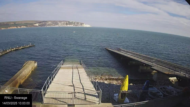 A view of a calm sea with a smooth surface reflecting the sky. In the foreground, there is a concrete ramp leading down to the water, bordered by a metal railing. To the right, a curved wooden pier juts out into the sea, while another pier extends straight ahead. On the ground near the ramp, there are yellow and blue kayaks. The background features a rocky shoreline and chalky cliffs under a clear blue sky with a few scattered clouds. The timestamp at the bottom indicates the image was taken on March 14, 2025, at 10:00 AM.