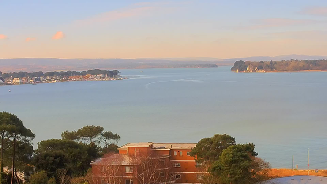 A panoramic view of a calm body of water under a clear blue sky with a few soft clouds. On the left side, there is a shoreline with buildings and boats, while on the right, the landscape features distant hills and a small island with trees. The foreground shows hints of greenery, including tree branches, along with a low-rise building. The overall scene conveys a peaceful and serene atmosphere.