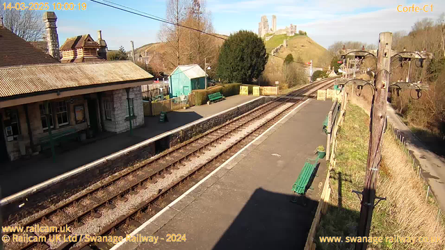 A view of Corfe Castle railway station on a clear day. The image shows a platform with a concrete surface, railway tracks extending in two directions, and several green benches along the platform. To the left, there is a building with a sloped roof and several windows. In the background, the ruins of Corfe Castle are prominent on a hill, surrounded by trees and grass. Utility poles are visible on the right side of the image, with wires extending overhead. The time stamp at the top indicates the date and time of the capture.