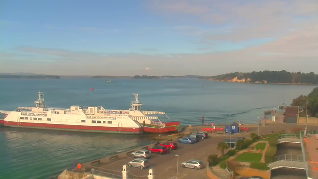 A view of a calm bay with a ferry docked at a terminal. The ferry is white with red accents, displaying a sign that instructs to give way to the chain ferry. In the foreground, there is a parking area with several cars, predominantly red and white, and a landscaped area with grass. The water is smooth, reflecting the blue sky, which has some clouds. In the distance, there are hills and trees along the shoreline.