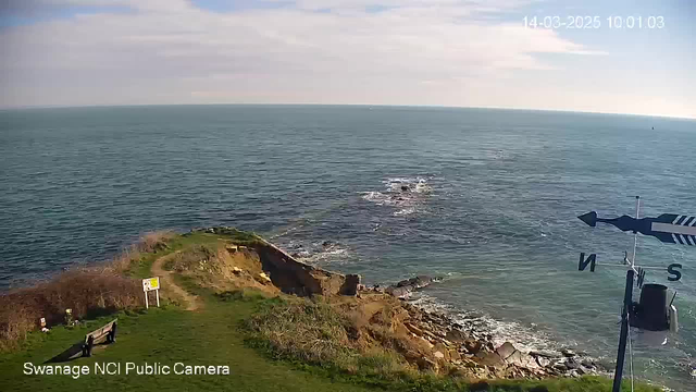 A coastal scene with a view of the ocean and rocky shoreline. Light blue water stretches out to the horizon under a partly cloudy sky. In the foreground, there is a grassy area with a bench and a sign. A weather vane showing the north direction is visible on the right side of the image. The overall atmosphere is calm and serene.