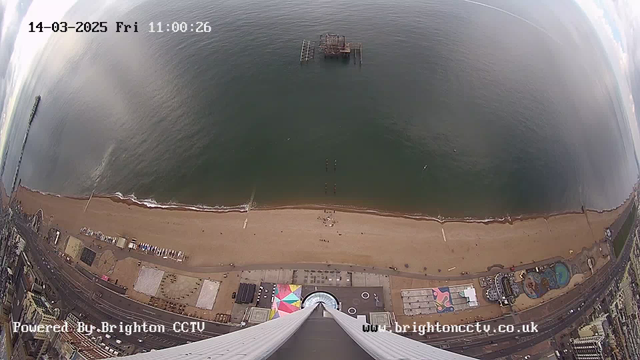 Aerial view of a beach and sea. The sandy beach extends along the bottom of the image, with a few people visible near the water's edge. Several structures, including a pier with an abandoned building, are situated in the water. The coastline is lined with buildings and attractions, with colorful patterns visible on some rooftops. The sky is partly cloudy, and there are gentle waves in the ocean. The image includes a timestamp at the top, indicating the date and time.