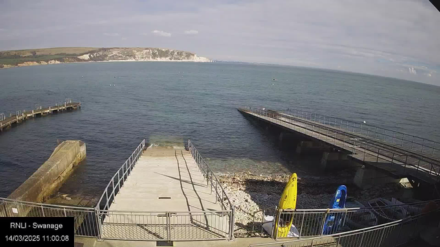 A waterfront scene showing a calm sea under a mostly clear sky. In the foreground, there are two docks: one with a smooth concrete surface leading to the water and the other with a wooden structure extending out into the sea. On the ground near the docks, there are rocks and small pebbles. To the right, there are two kayaks, one yellow and one blue, positioned on the shore. The background features a gentle incline towards green hills and white cliffs along the coastline.