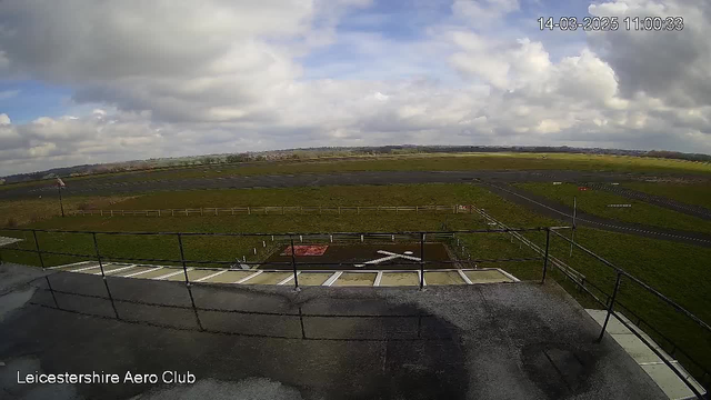 A landscape view from a high vantage point at Leicestershire Aero Club. The foreground shows a flat rooftop area with a railing and some markings. The midground features an airstrip with a few dark pavement sections and a grassy area in between. There are some wooden fences and a few signs, while the distant background reveals a line of trees and clouds in a partly cloudy sky. The overall scene is open and expansive, featuring a mix of paved and grassy surfaces under a bright sky.