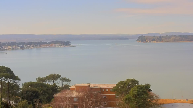 A serene view of a calm body of water, with distant hills under a clear sky. In the foreground, a building made of reddish bricks is partially visible, surrounded by green trees. The water reflects the soft colors of the sky, and there are several boats visible in the distance near a marina. The shoreline features various structures, including what appears to be a lighthouse or similar building on the right.