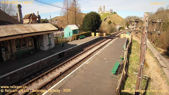 A railway station platform viewed from an elevated angle. The platform is lined with green benches and a small blue shed on the left. Stone walls surround sections of the platform. In the background, a castle ruins sits atop a green hill under a clear blue sky. Train tracks run parallel to the platform, leading off into the distance. A wooden pole with wires can be seen on the right side of the image.